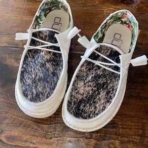 May include: A pair of white leather shoes with a black and white cowhide pattern. The shoes have a floral print lining and the text "The  CU" is visible on the inside of the shoes.