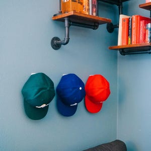 May include: Three baseball caps in solid dark green, royal blue, and bright red are mounted on a blue wall. Above the caps, industrial-style pipe shelving holds various books, a red candle, a round clock, and a vintage red camera.