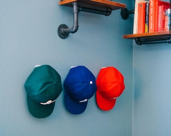 May include: Three baseball caps in solid dark green, royal blue, and bright red are mounted on a blue wall. Above the caps, industrial-style pipe shelving holds various books, a red candle, a round clock, and a vintage red camera.