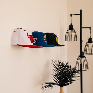 May include: Three baseball caps mounted on a wall. The first is white with a red brim and a red bull logo. The second is blue with a yellow and blue circular emblem. The third is black with a yellow circular logo featuring a stylized figure.
