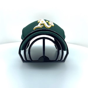 May include: A dark green baseball cap featuring the white and yellow embroidered letters A's on the front. The cap is resting on a black plastic curved display stand that holds the shape of the crown and the brim in a rounded position.