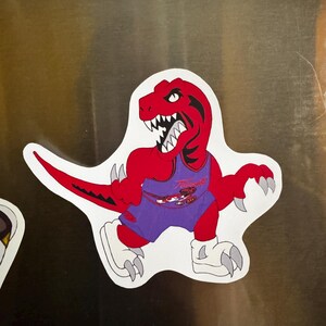 Peut inclure: Autocollant découpé représentant un dinosaure rouge de dessin animé portant un maillot violet avec le mot "Raptors". Le dinosaure a des dents blanches acérées, des griffes grises et des chaussures blanches. L'autocollant est sur une surface métallique.