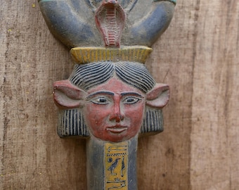 May include: A wooden sculpture of an Egyptian goddess with a cobra on her head. The sculpture is painted in red, blue, yellow, and black. The goddess is wearing a crown with a cobra on top. The sculpture is standing on a wooden base.