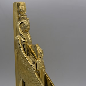 Może przedstawiać: Złota statua egipskiej bogini Izydy z skrzydłami, trzymająca małą figurkę faraona. Statua stoi na podstawie z hieroglifami.