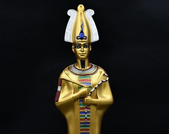 May include: A gold statue of an Egyptian pharaoh, standing on a rectangular base. The pharaoh wears a headdress with white and gold details, and holds a staff. The statue has colourful details and is set against a black background.