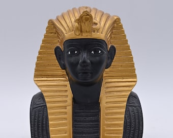 Könnte beinhalten: Eine schwarze und goldene Figur eines ägyptischen Pharaos, der eine traditionelle Kopfbedeckung mit einem Kobramuster trägt. Das Gesicht des Pharaos ist mit schwarzen Augen und einem ernsten Ausdruck detailliert.