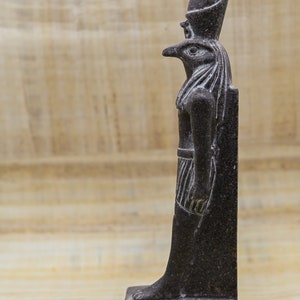 Puede incluir: Una estatua de piedra de color gris oscuro que representa al dios egipcio Horus, representado como un hombre con cabeza de halc&oacute;n y una corona alta. La estatua est&aacute; sobre una base con jerogl&iacute;ficos.