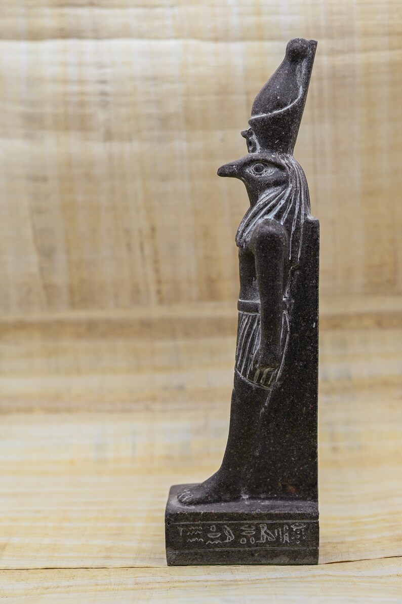Puede incluir: Una estatua de piedra de color gris oscuro que representa al dios egipcio Horus, representado como un hombre con cabeza de halc&oacute;n y una corona alta. La estatua est&aacute; sobre una base con jerogl&iacute;ficos.