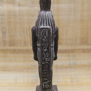 Puede incluir: Estatua de piedra negra de un dios egipcio con jerogl&iacute;ficos tallados en la parte delantera. La estatua est&aacute; de pie sobre una base rectangular.