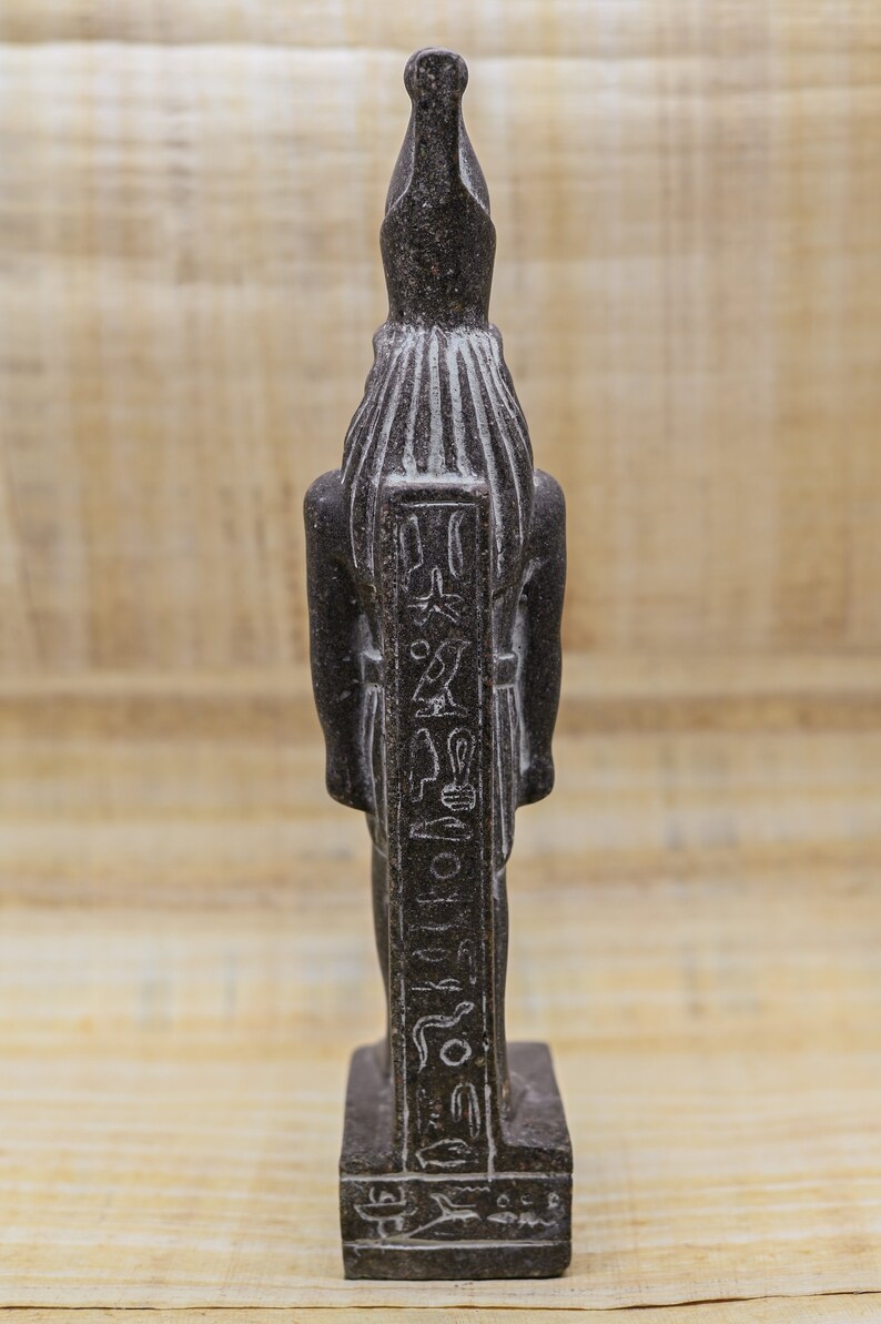 Puede incluir: Estatua de piedra negra de un dios egipcio con jerogl&iacute;ficos tallados en la parte delantera. La estatua est&aacute; de pie sobre una base rectangular.
