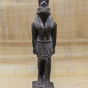 Puede incluir: Una estatua de piedra gris oscura de un dios egipcio con un tocado y una falda. La estatua est&aacute; de pie sobre una base rectangular con jerogl&iacute;ficos.