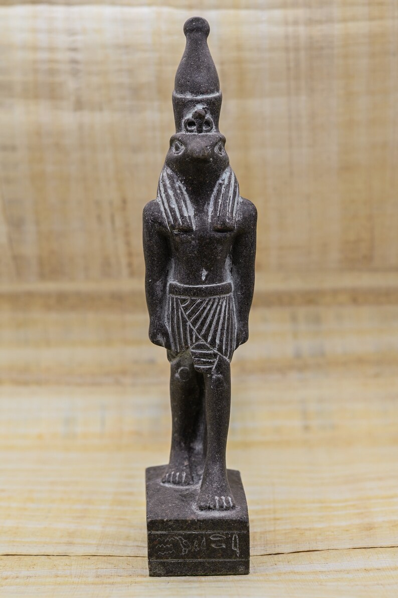 Puede incluir: Una estatua de piedra gris oscura de un dios egipcio con un tocado y una falda. La estatua est&aacute; de pie sobre una base rectangular con jerogl&iacute;ficos.