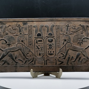 May include: A rectangular stone relief with a detailed carving of two Egyptian deities. Each deity is depicted as a lion with a human head wearing a crown. The deities are flanking a central cartouche with hieroglyphs. The relief is brown and black.