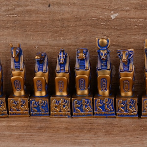 May include: A set of nine gold and blue Egyptian god statues. Each statue is a different god with a unique head and body. The statues are all standing on a wooden surface.