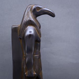 Puede incluir: Una estatua negra y dorada del dios egipcio Horus, representado como un hombre con cabeza de halcón. La estatua está de pie sobre una base rectangular.