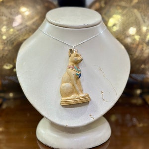 Może przedstawiać: Złoty naszyjnik z detalicznym, egipskim kotkiem w formie zawieszki, z niebieskimi i czerwonymi akcentami. Naszyjnik jest prezentowany na białym manekinie.