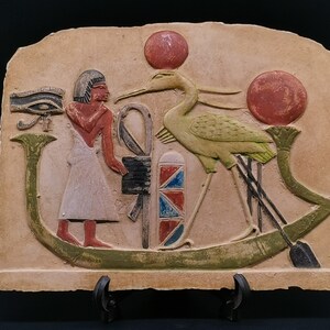 May include: A beige stone relief depicting an Egyptian god with a white robe and a green bird with red circles above it. The god is holding a staff and a symbol with red, white, and blue stripes. The relief is mounted on a black stand.