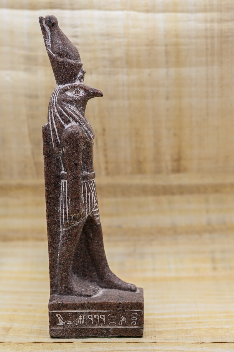 Puede incluir: Una estatua de granito marr&oacute;n del dios egipcio Horus, representado como un hombre con cabeza de halc&oacute;n. La estatua est&aacute; de pie sobre una base con jerogl&iacute;ficos que dicen "Horus, el Grande".
