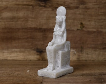 May include: White ceramic figurine of the Egyptian goddess Bastet, depicted as a seated lioness with a human head and a crown. The figurine is detailed with intricate carvings and is sitting on a small base.