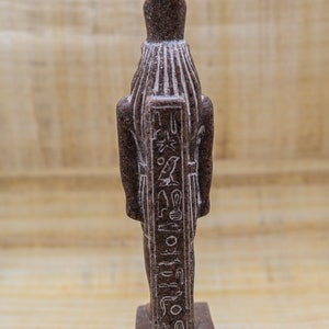Puede incluir: Una peque&ntilde;a estatua de piedra marr&oacute;n de un dios egipcio con jerogl&iacute;ficos tallados en la parte delantera. La estatua est&aacute; de pie sobre una peque&ntilde;a base.