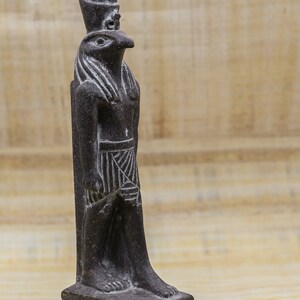 Puede incluir: Una estatua de piedra de color gris oscuro que representa al dios egipcio Horus, con cuerpo humano y cabeza de halc&oacute;n. La estatua est&aacute; sobre una base rectangular adornada con jerogl&iacute;ficos.