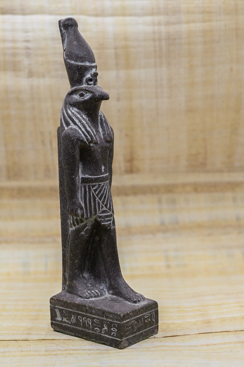 Puede incluir: Una estatua de piedra de color gris oscuro que representa al dios egipcio Horus, con cuerpo humano y cabeza de halc&oacute;n. La estatua est&aacute; sobre una base rectangular adornada con jerogl&iacute;ficos.