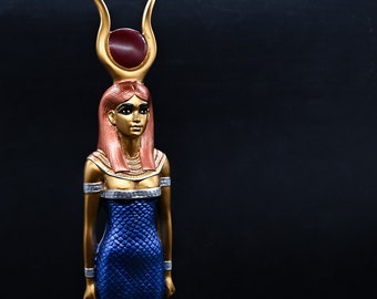 May include: A gold statue of an Egyptian goddess with a red gem in her crown and a blue and gold dress.