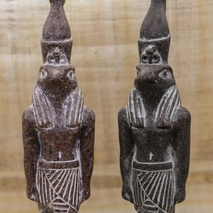 Puede incluir: Dos peque&ntilde;as estatuas de color marr&oacute;n oscuro del dios egipcio Anubis, cada una con cabeza de chacal y cuerpo humano. Las estatuas est&aacute;n de pie y tienen una superficie detallada y texturizada.