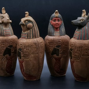 Op de afbeelding: Vier decoratieve canopische potten met Egyptisch geïnspireerde deksels. De deksels tonen een ram, een vogel, een vrouw met een hoofdtooi en een jakhals. De potten zijn bruin met hiërogliefenontwerpen.