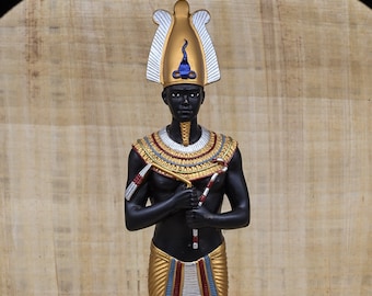 May include: A black statue of an Egyptian pharaoh wearing a gold and white headdress with a blue and gold cobra design. The pharaoh is holding a staff in his right hand and a crook in his left hand. The statue is standing on a beige background.