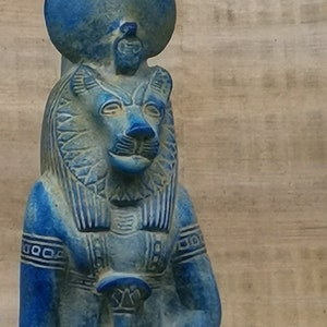 May include: Blue and white statue of an Egyptian goddess with a lion head and a crown. The statue is carved with intricate details and has a weathered look.