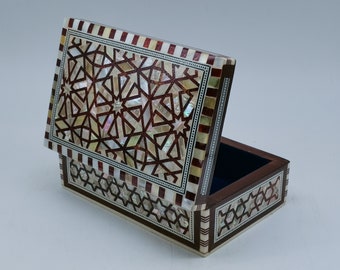 Puede incluir: Una pequeña caja de madera con tapa abatible, decorada con intrincados patrones geométricos hechos de incrustaciones de nácar. La caja tiene una base de madera marrón oscuro y un borde a rayas rojo y blanco.