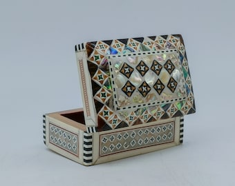 Puede incluir: Una pequeña caja rectangular de madera con un intrincado diseño de incrustaciones. La caja está hecha de madera y nácar, y presenta un patrón geométrico en tonos marrones, blancos y verdes. La tapa está abierta, revelando el interior de la caja.