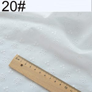 May include: White fabric featuring a repeating embroidered floral pattern with small eyelet details. A wooden ruler is positioned across the bottom left corner, showing a scale of 11 centimeters to provide a clear sense of the fabric scale.
