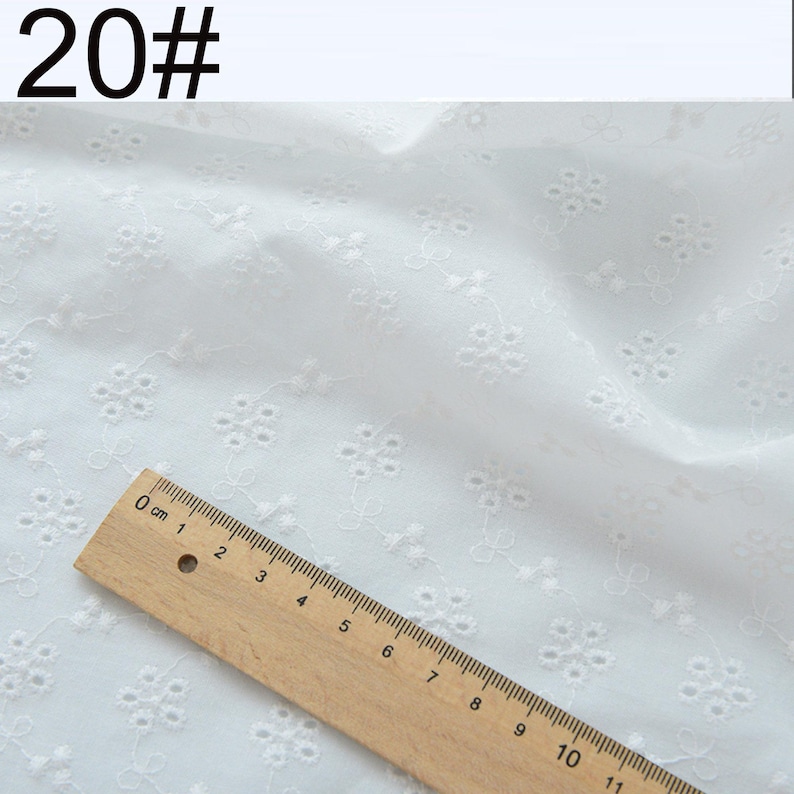 May include: White fabric featuring a repeating embroidered floral pattern with small eyelet details. A wooden ruler is positioned across the bottom left corner, showing a scale of 11 centimeters to provide a clear sense of the fabric scale.