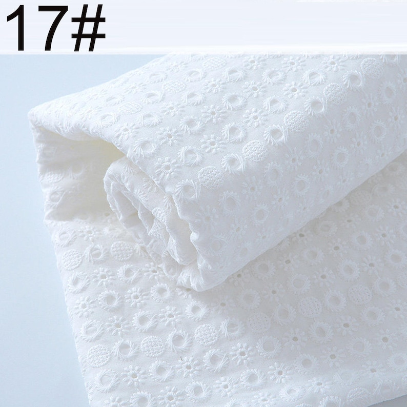 May include: A folded piece of white eyelet fabric featuring a repeating pattern of small embroidered flowers and circular cutouts. The fabric is labeled with the text 17# in the upper left corner, showcasing a delicate and intricate texture.