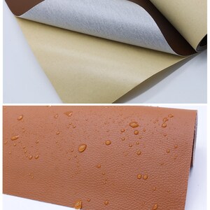 May include: Close-up of a brown leather-like fabric with a textured surface. Water droplets are visible on the fabric, demonstrating its water-resistant properties. The fabric has a backing layer with adhesive for easy application.