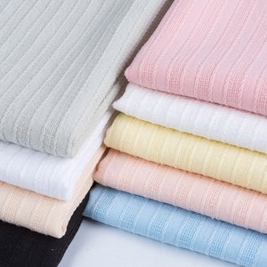 K&ouml;nnte beinhalten: Ein Stapel Stoffmuster in verschiedenen Farben, darunter Grau, Rosa, Wei&szlig;, Gelb, Pfirsich, Blau und Schwarz. Der Stoff hat eine strukturierte, gerippte Oberfl&auml;che.