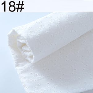 May include: A roll of white fabric featuring an intricate eyelet embroidery pattern with small circular cutouts and delicate floral vine motifs. The textile is folded over itself, displaying the consistent texture and the text 18# in the corner.