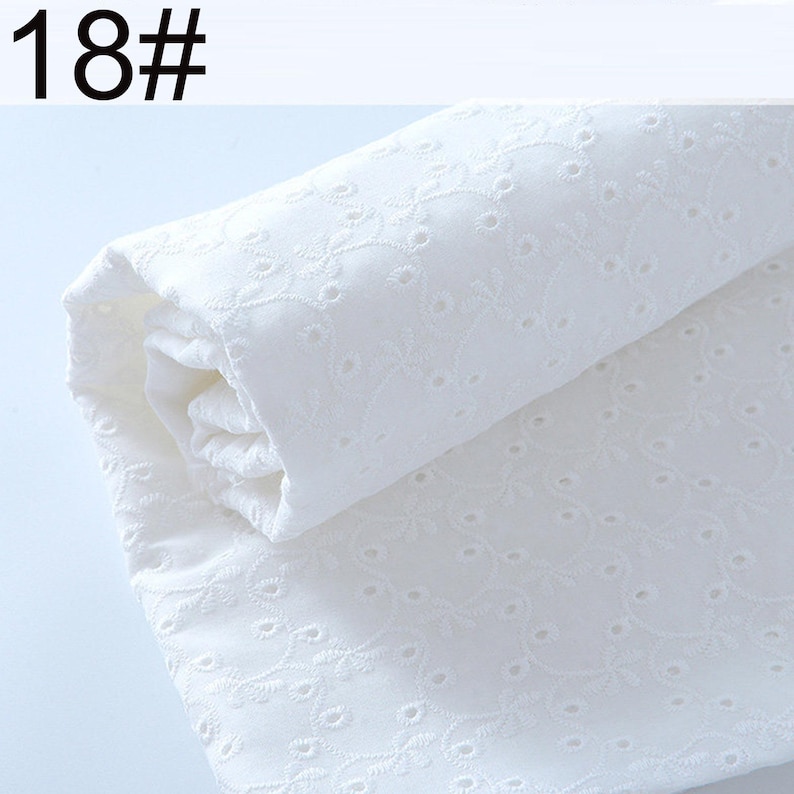 May include: A roll of white fabric featuring an intricate eyelet embroidery pattern with small circular cutouts and delicate floral vine motifs. The textile is folded over itself, displaying the consistent texture and the text 18# in the corner.
