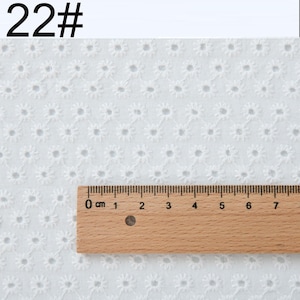 May include: A white eyelet fabric featuring a repeating floral pattern with small circular cutouts. A wooden ruler is placed horizontally across the fabric, showing a scale from 0 to 7 inches. The text 22# is visible in the top left corner.
