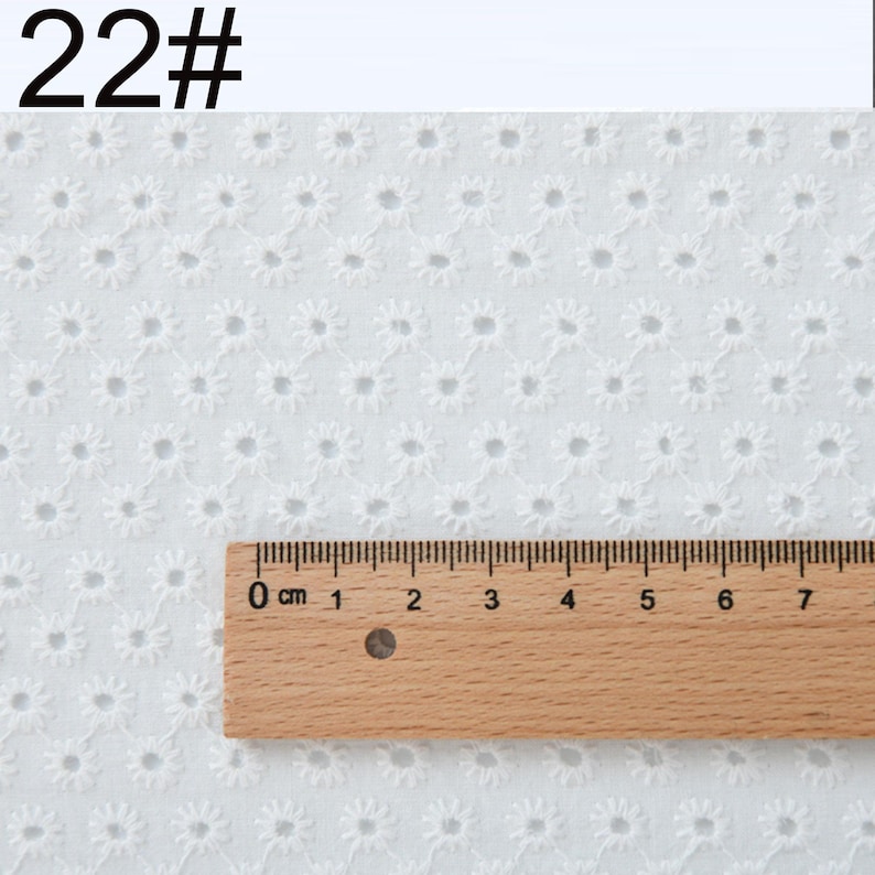 May include: A white eyelet fabric featuring a repeating floral pattern with small circular cutouts. A wooden ruler is placed horizontally across the fabric, showing a scale from 0 to 7 inches. The text 22# is visible in the top left corner.