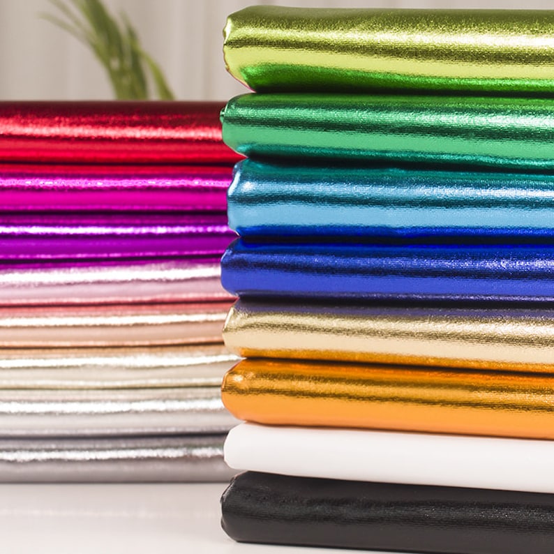 Op de afbeelding: Een stapel metallic stof in verschillende kleuren, waaronder goud, zilver, zwart, wit, oranje, blauw, groen, roze en rood.