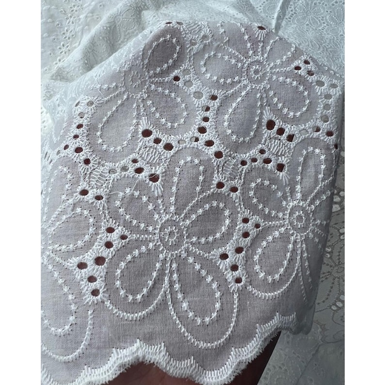May include: A white fabric featuring an intricate eyelet embroidery pattern with repeating floral motifs. The material displays a scalloped edge and a series of small, circular cutouts arranged in a decorative design across the surface.