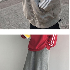 May include: A gray sweatshirt with white stripes on the sleeves. The sweatshirt has a zipper closure and a high neckline. The model is also wearing gray sweatpants with white stripes on the sides.