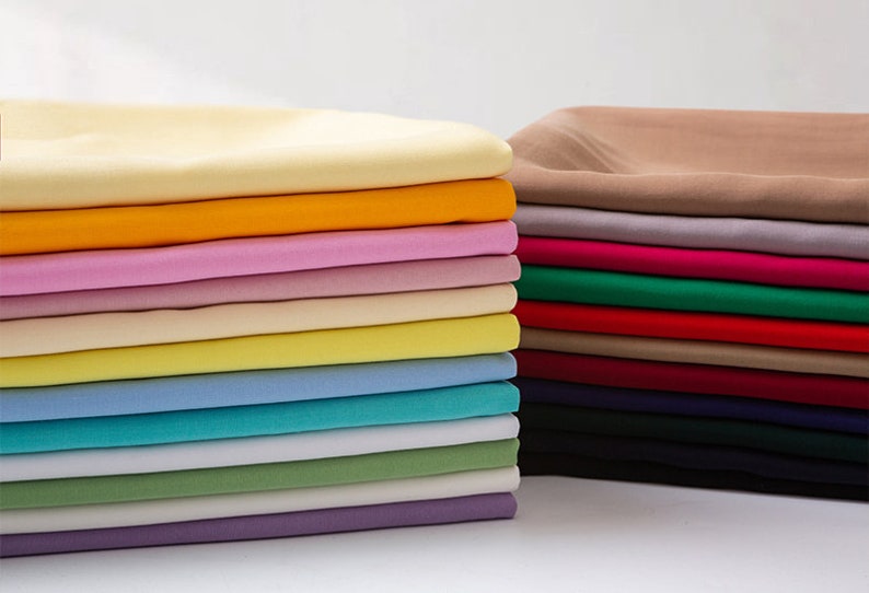 K&ouml;nnte beinhalten: Ein Stapel von 18 Stoffproben in verschiedenen Farben, darunter Beige, Gelb, Rosa, Hellblau, Gr&uuml;n und Lila. Die Proben sind in einem ordentlichen Stapel angeordnet, wobei die oberste Probe beige ist.