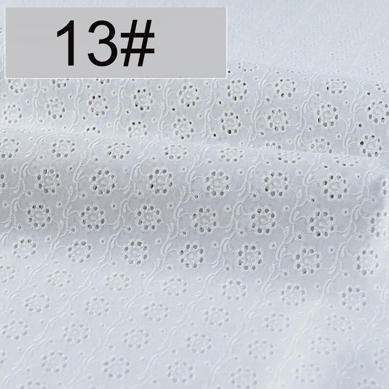 May include: A white fabric featuring an eyelet embroidery pattern with repeating floral motifs and delicate swirling vine details. The textile displays a consistent perforated design throughout, and a label marked 13# is visible.