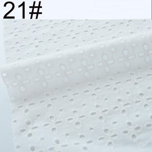 May include: A white fabric featuring an eyelet embroidery pattern with repeating floral motifs. The textile displays a series of small, circular cutouts arranged in four-petal flower shapes across the surface. The text 21# is visible.