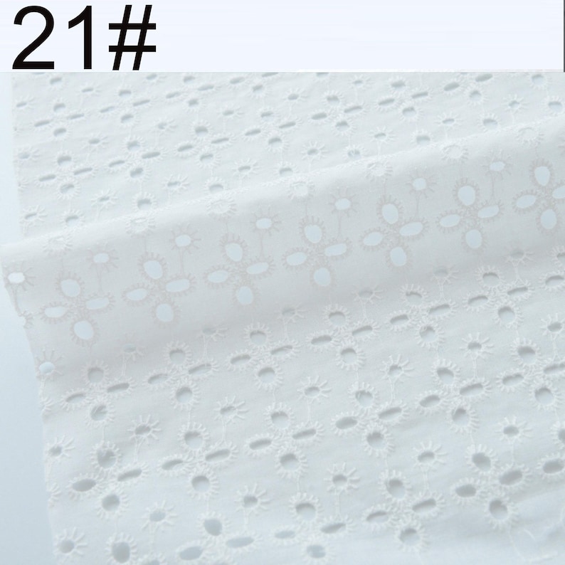 May include: A white fabric featuring an eyelet embroidery pattern with repeating floral motifs. The textile displays a series of small, circular cutouts arranged in four-petal flower shapes across the surface. The text 21# is visible.
