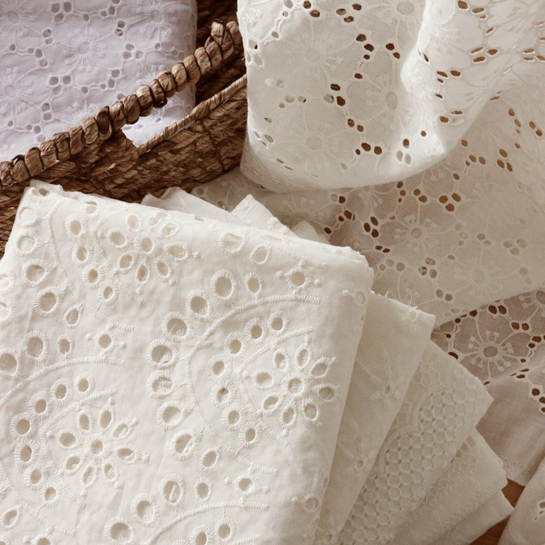 May include: A stack of folded white cotton fabric featuring intricate eyelet embroidery with floral patterns and circular cutouts. The fabric is layered and partially draped, showcasing the detailed needlework and the texture of the material.